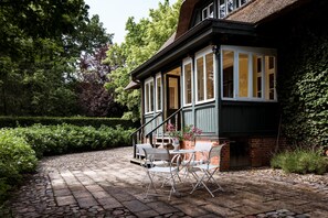 A covered patio with a brick floor, white outdoor furniture, and a view of a house with a green roof and white-framed windows. - Wunderschönes Landhaus (Aumuehle)