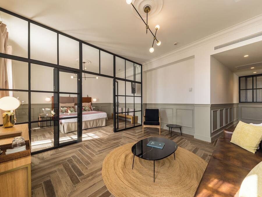Una habitación de hotel moderna con un amplio ventanal corredero que da al dormitorio, un tapete redondo tejido, una mesa de centro negra y un sofá de cuero.