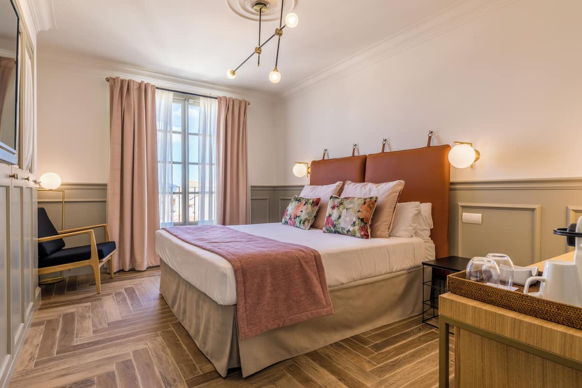 A hotel room with a large bed, a wooden floor, a desk with a kettle, and a window with curtains.