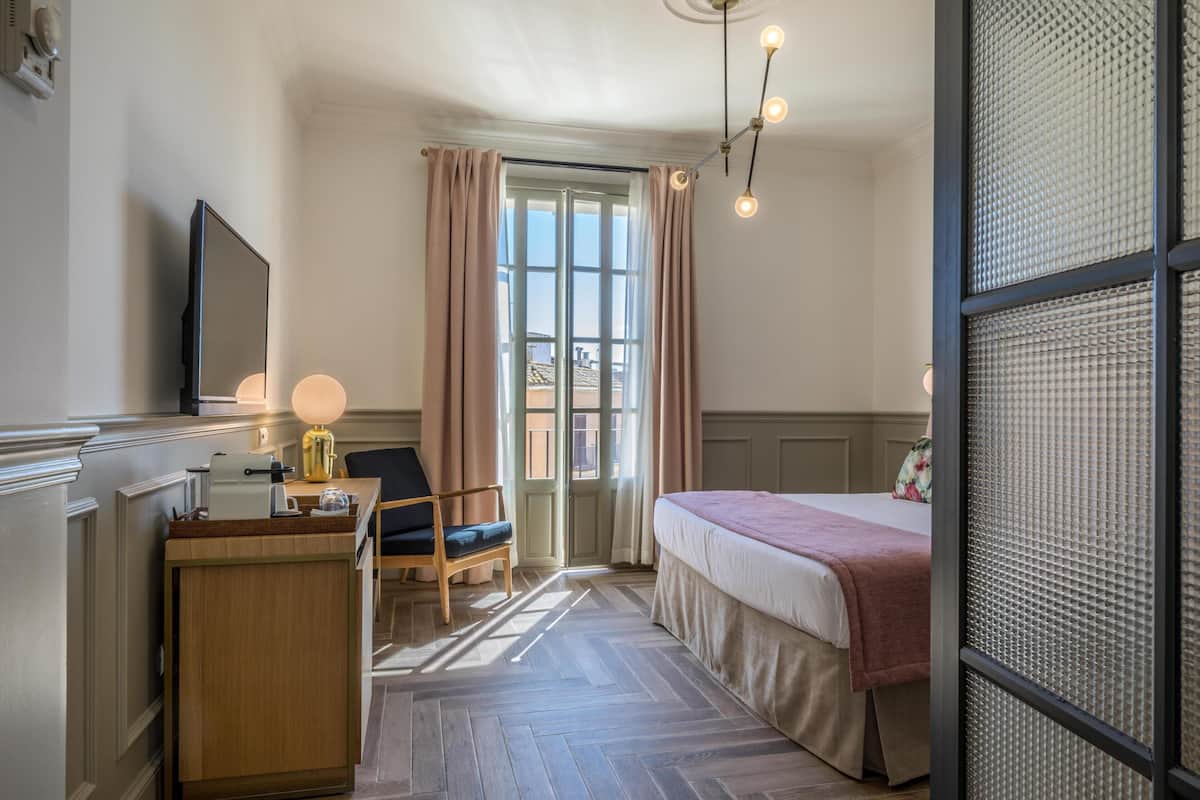 A hotel room with a bed, a desk with a lamp, a chair, and a large window with curtains.
