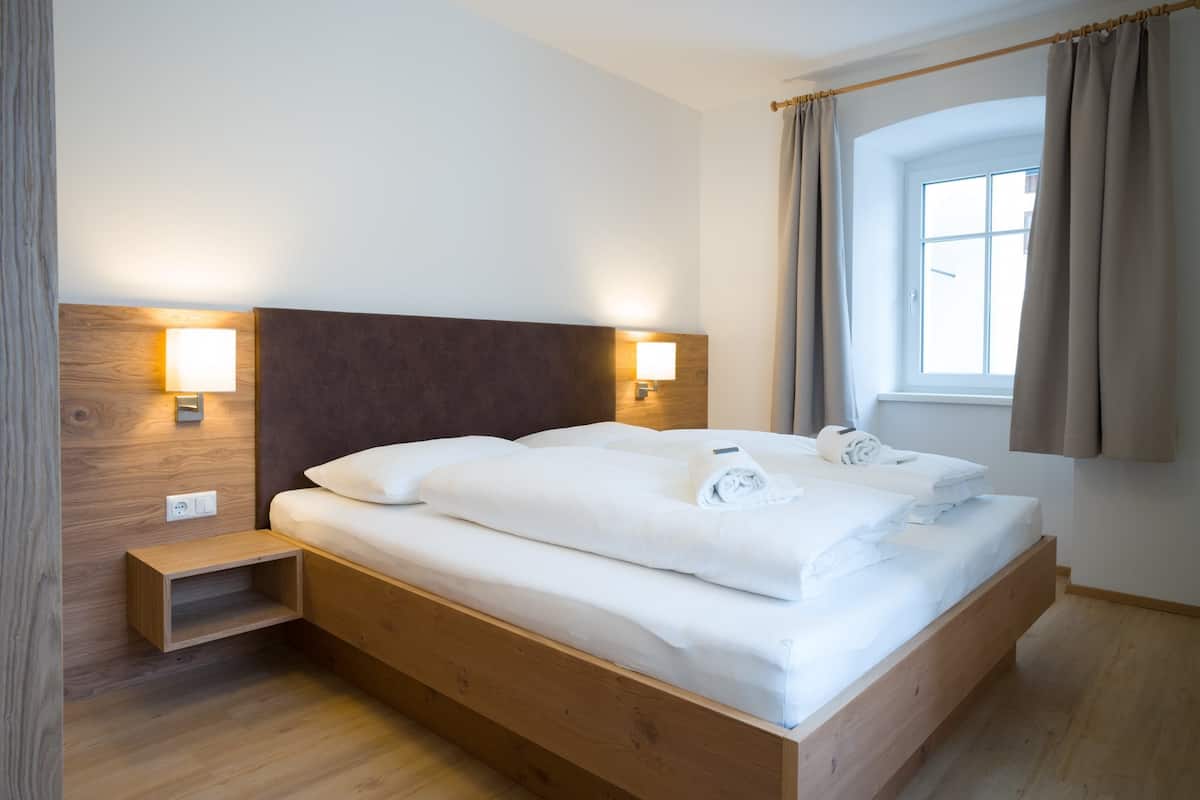 A modern hotel room with a wooden bed, bedside lamps, a window with curtains, and a small wooden nightstand.
