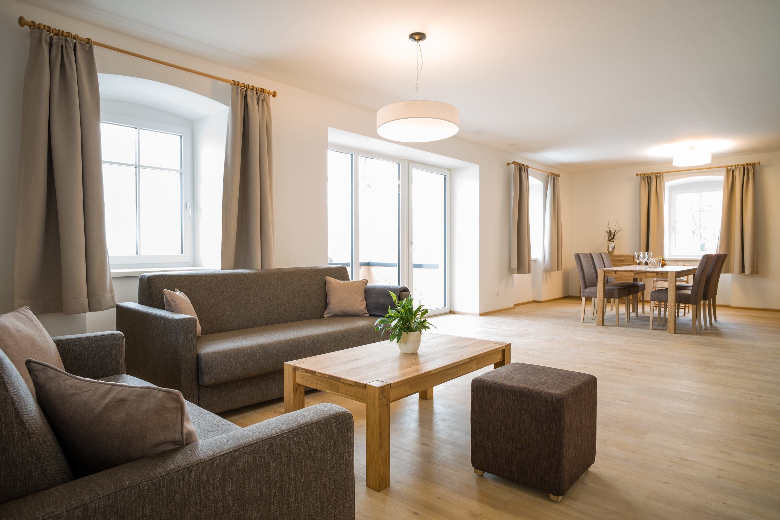 A modern living room with a sofa, ottoman, and coffee table, a dining area with a table and chairs, and large windows with curtains.