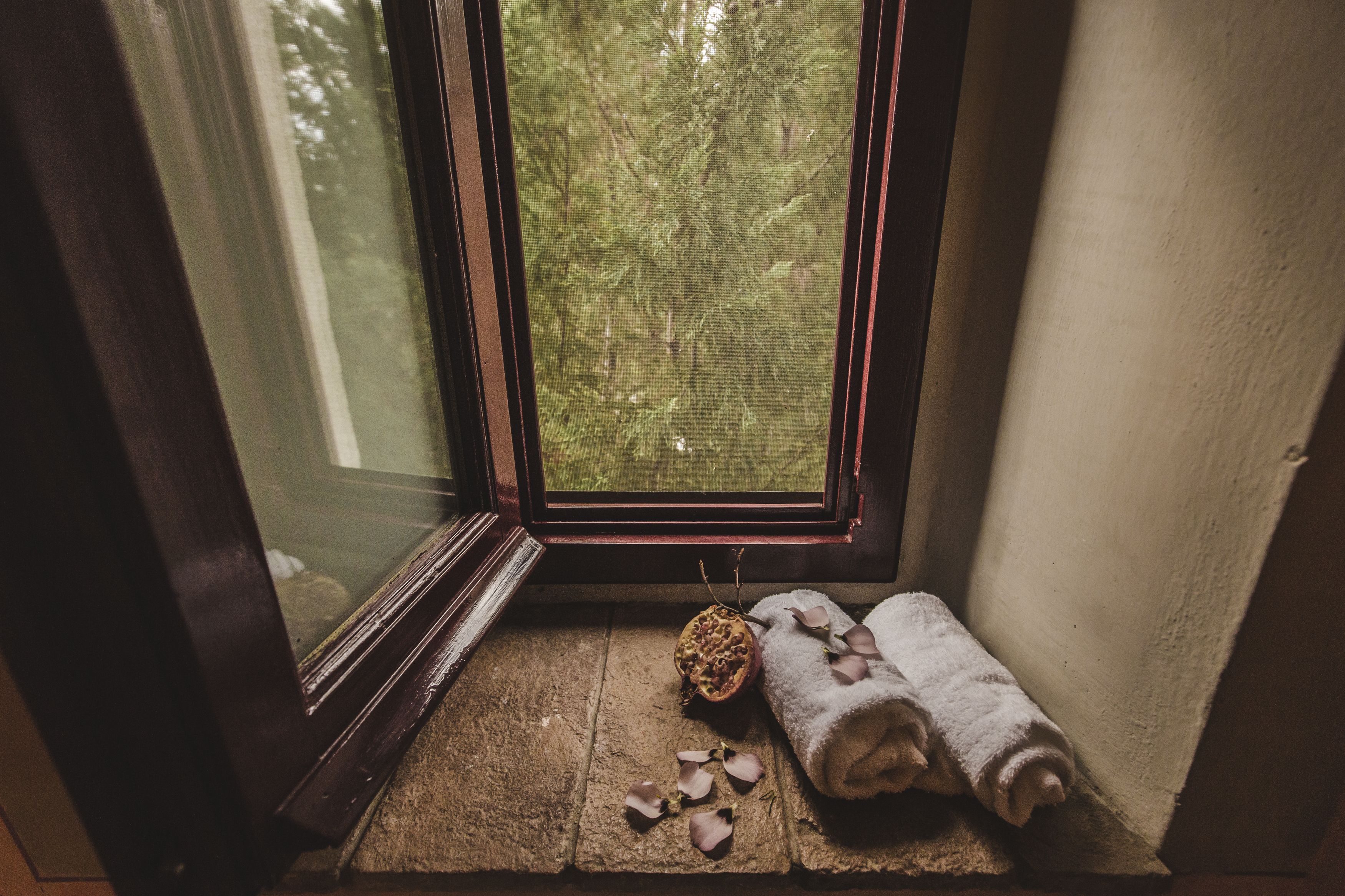 A small window with a view of a forest, a stone floor, a pomegranate, and a rolled towel with rose petals.