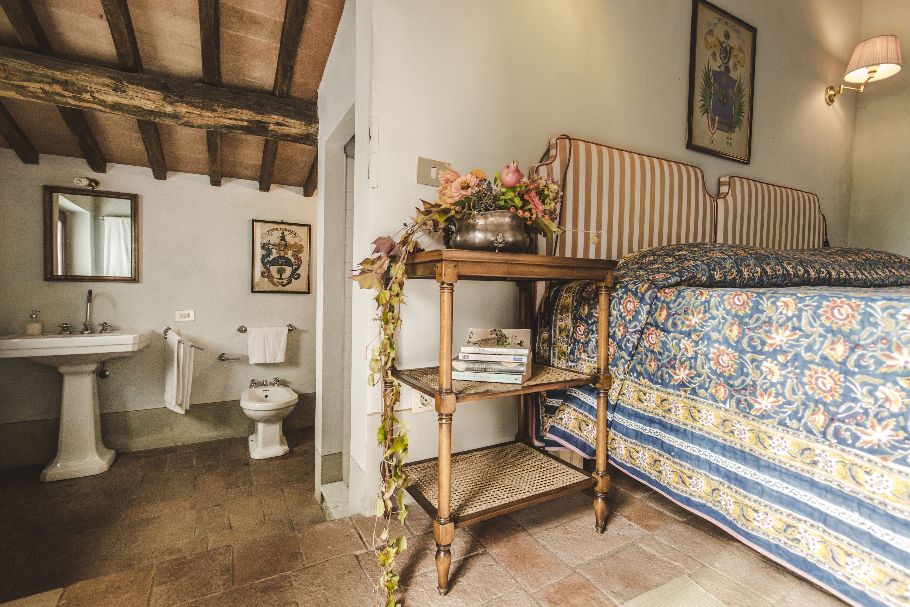 Quarto com cama, mesa de cabeceira com um vaso de flores, cabeceira de madeira, luminária na parede e um quadro emoldurado acima da cama.