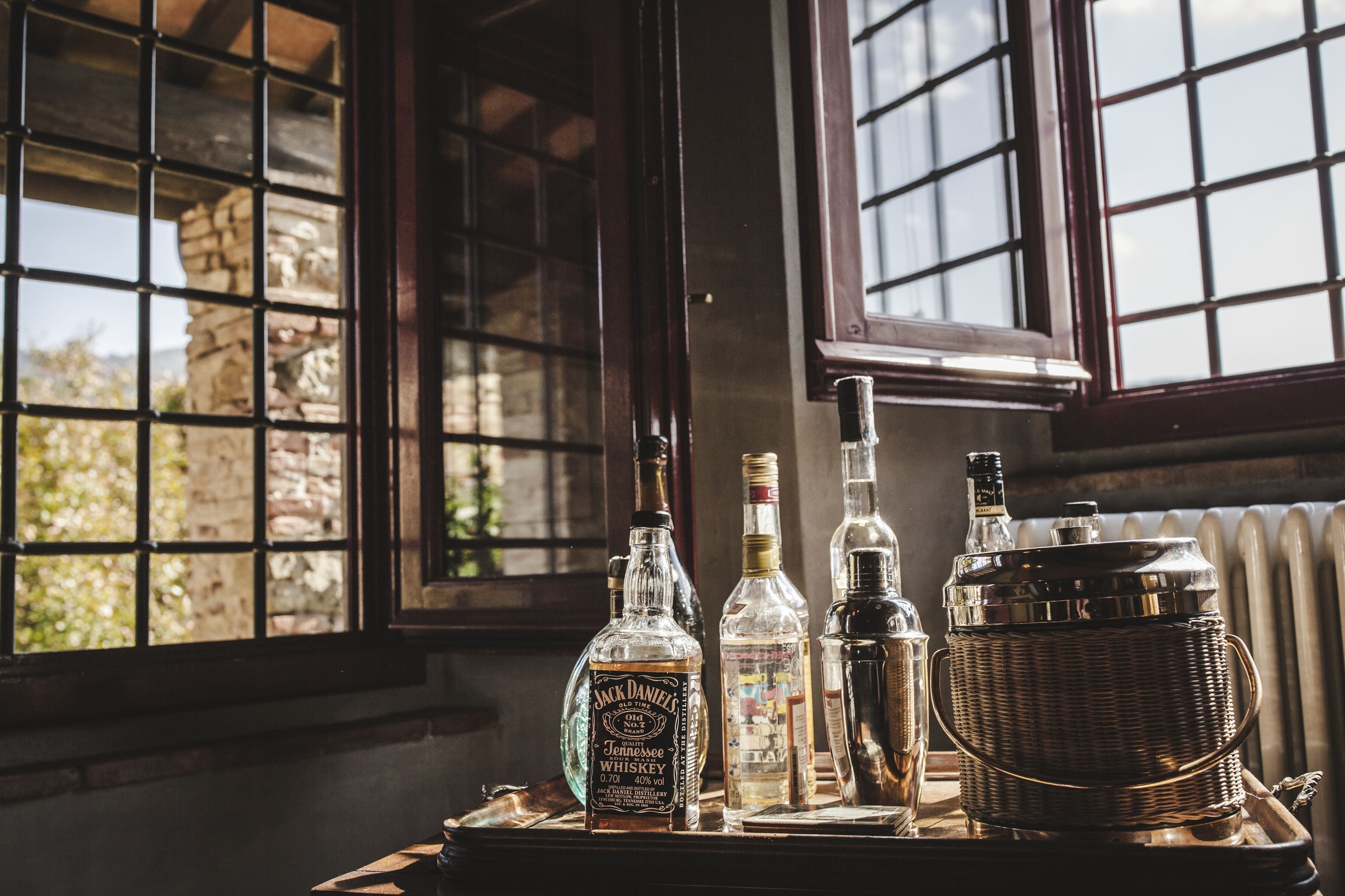 A table with various bottles and a metal container, likely for serving drinks, near a window with a view of greenery.