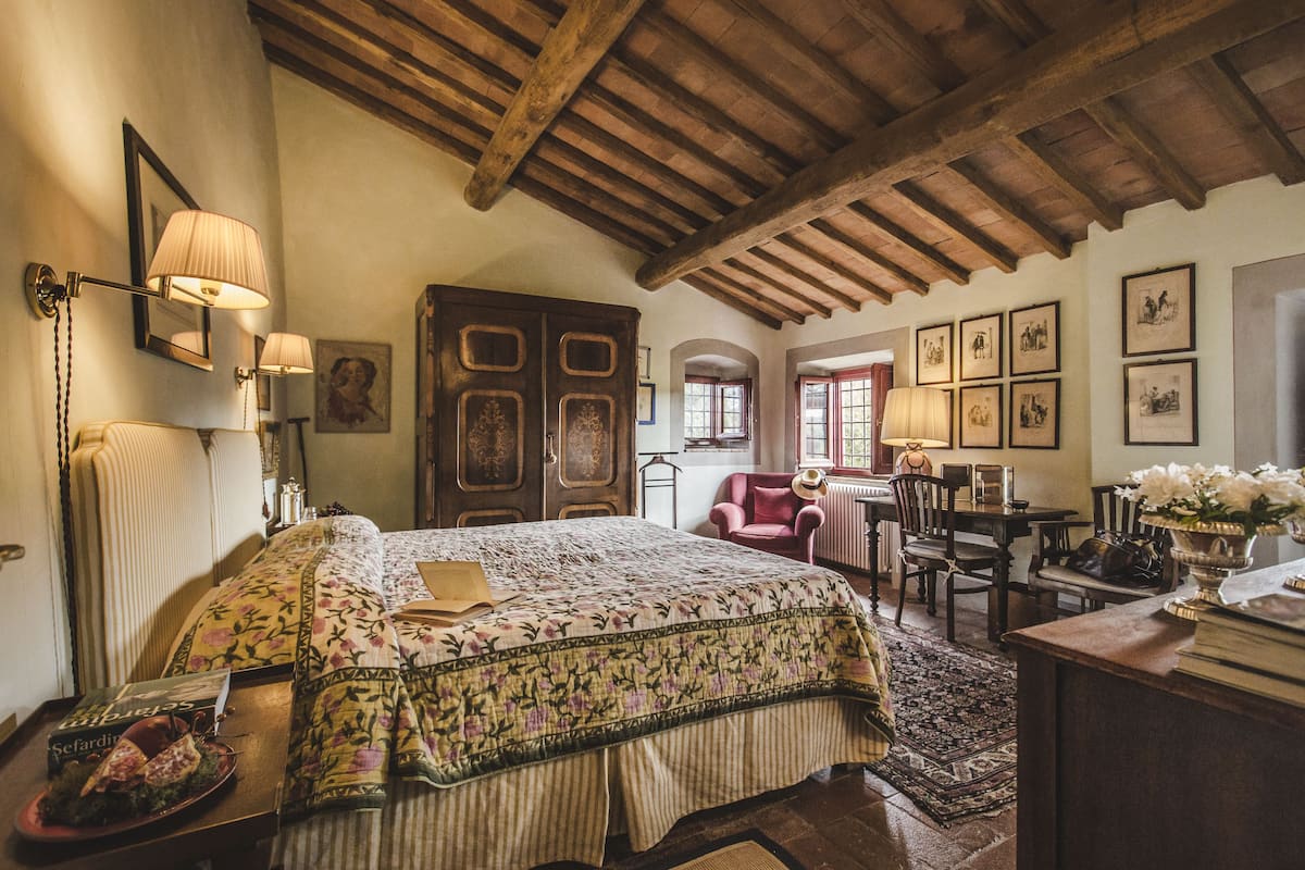 A traditional bedroom with a bed, a desk, and a chair. There are framed pictures on the wall and a lamp on the desk.