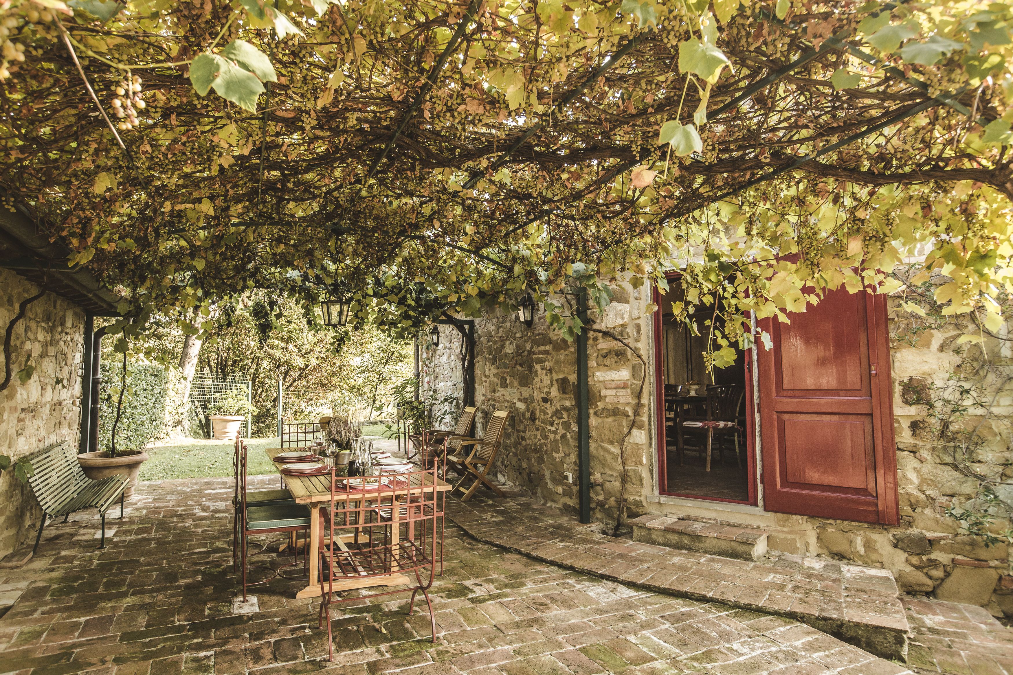 Un'area salotto esterna rustica con un muro in pietra, un tavolo di legno e sedie, immersa nel verde e con una porta rossa.
