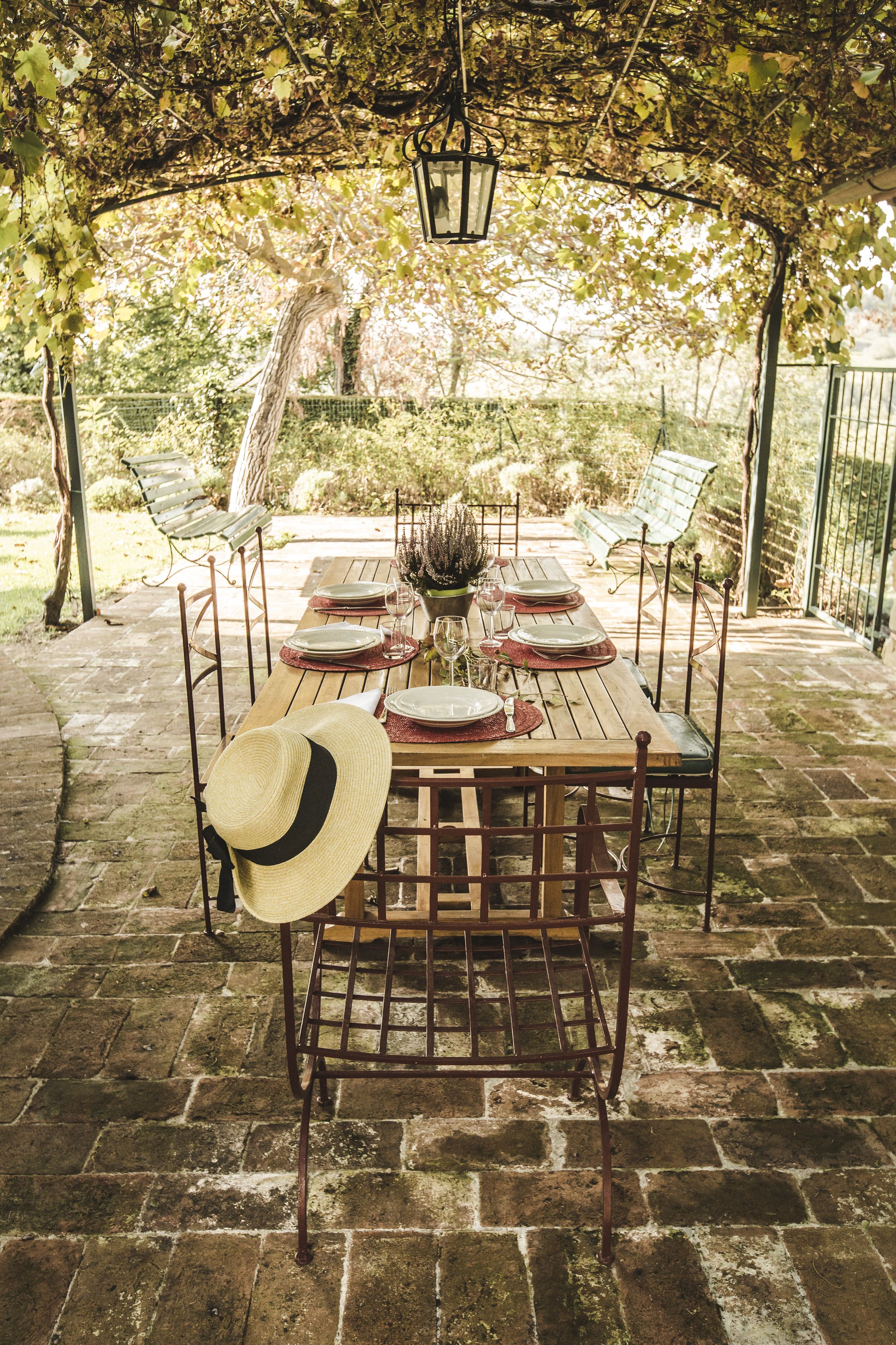 Mesa de madera al aire libre dispuesta para comer, rodeada de sillas de metal, bajo un área cubierta con un farol colgante.