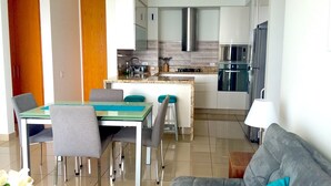 A modern kitchen with stainless steel appliances, a dining area with a glass table and grey chairs, and a grey sofa in the living area. - H2 LUXURY 3 Bedroom Apartment (Cartagena)