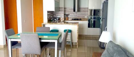 Una cocina moderna con electrodomésticos de acero inoxidable, una zona de comedor con mesa de vidrio y sillas grises, y un sofá gris en la sala de estar.