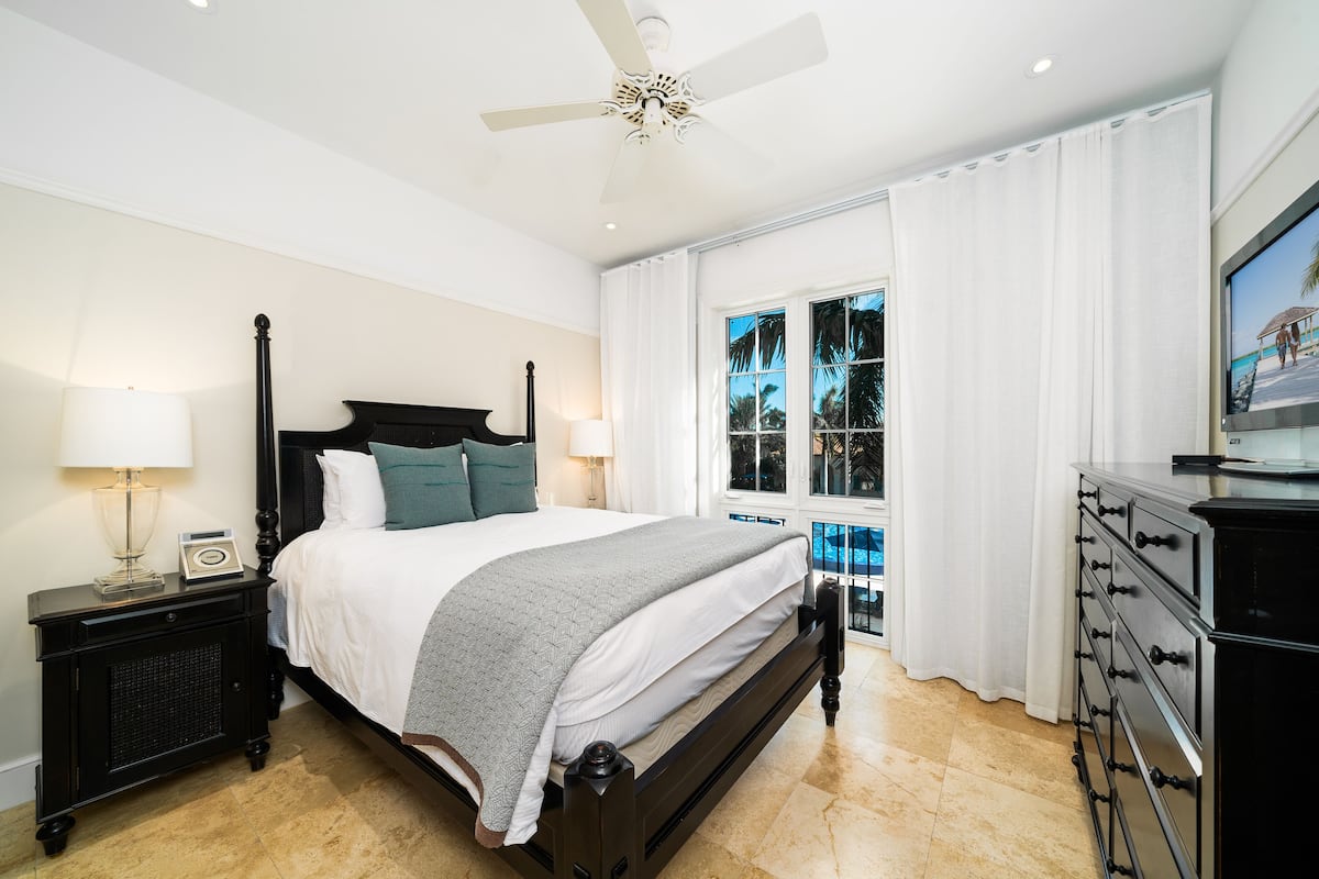 A bedroom with a four-poster bed, a nightstand with a lamp, a dresser, and a large window with curtains.