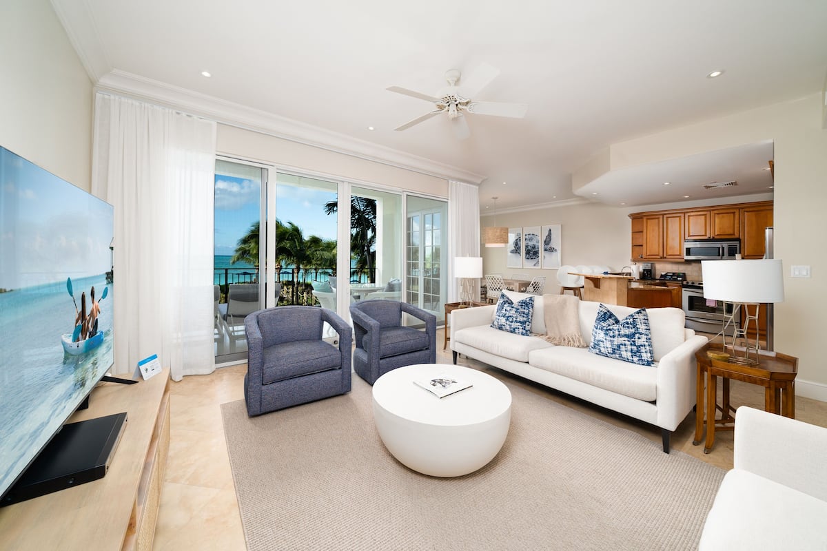 A spacious living room with a flat-screen TV, two armchairs, a white sofa, and a round coffee table. A ceiling fan and a kitchen area are visible in the background.