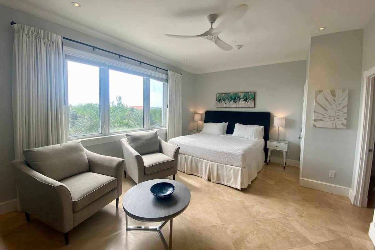 A modern hotel room with a bed, two armchairs, a small table, a ceiling fan, and large windows.