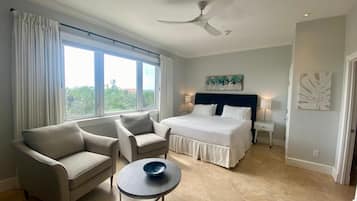 A modern hotel room with a bed, two armchairs, a small table, a ceiling fan, and large windows.
