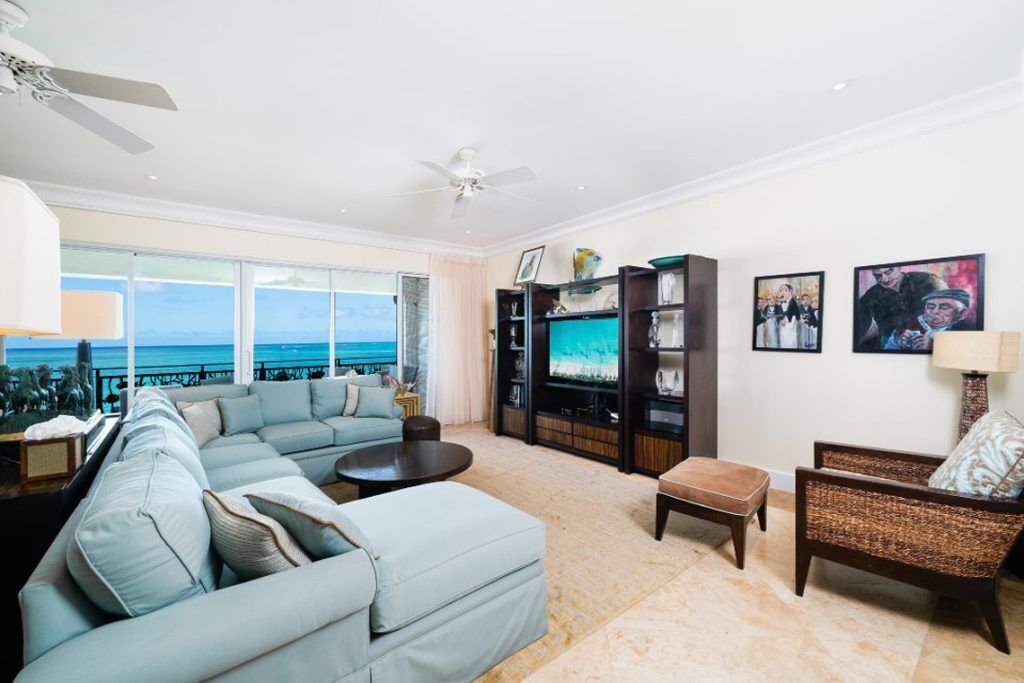 A spacious living room with a large sofa, a coffee table, and a television. There are two chairs and a small table. The room has a view of the ocean.