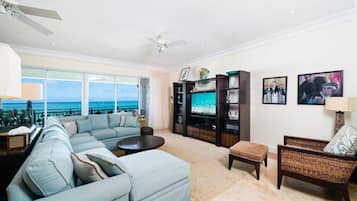 A spacious living room with a large sofa, a coffee table, and a television. There are two chairs and a small table. The room has a view of the ocean.