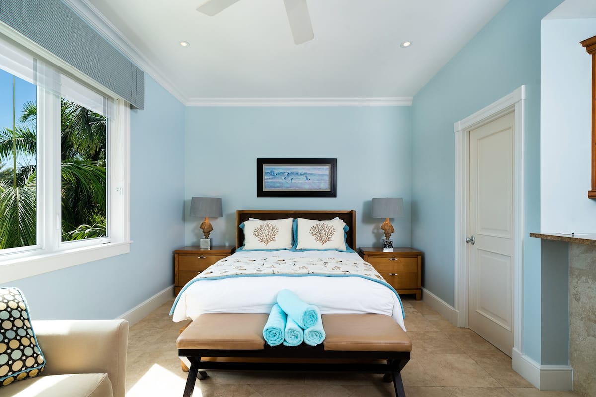 A bedroom with a bed, nightstands, a bench, and a window with a view of palm trees.