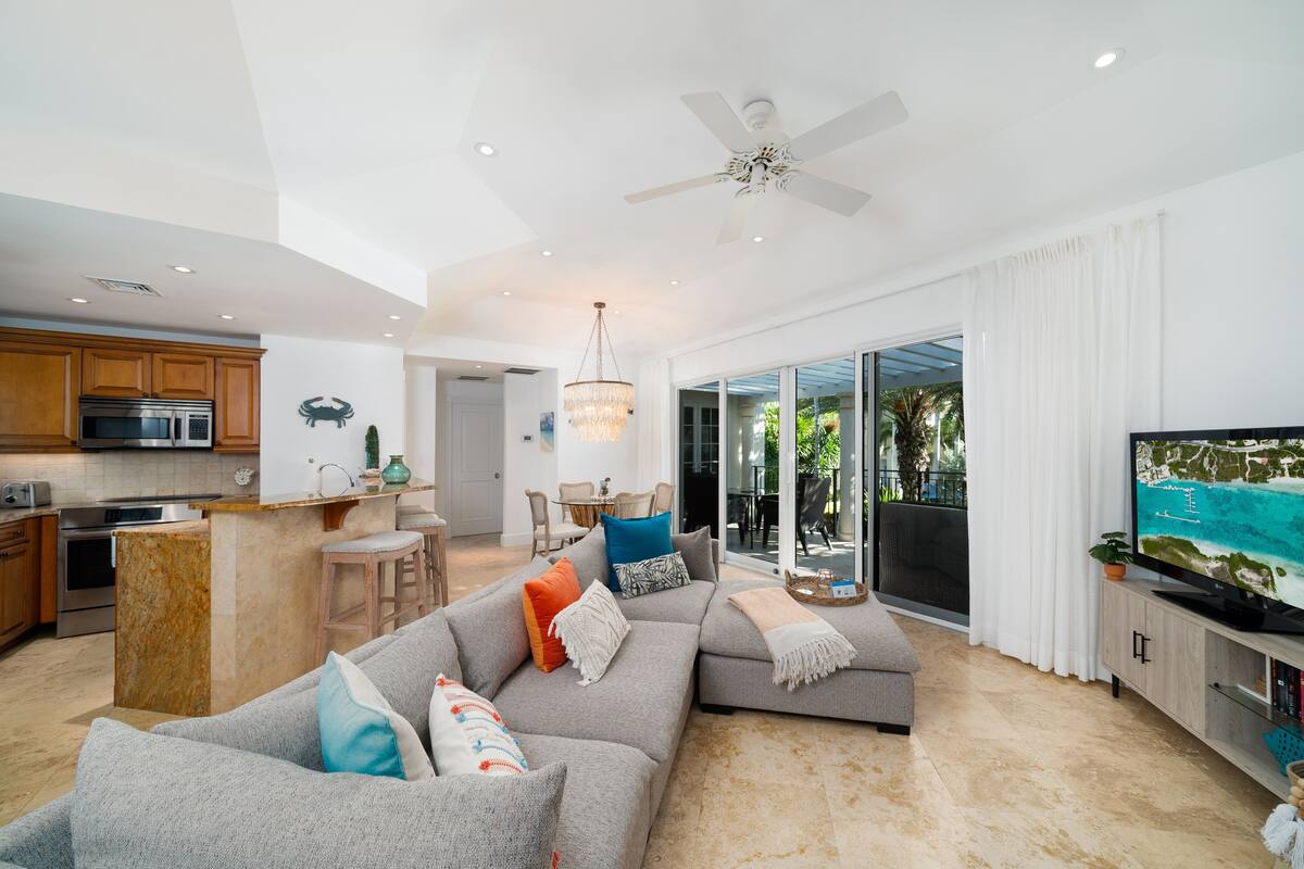 A modern living room with a grey sectional sofa, a kitchen with wooden cabinets, and a large TV on a wooden cabinet.