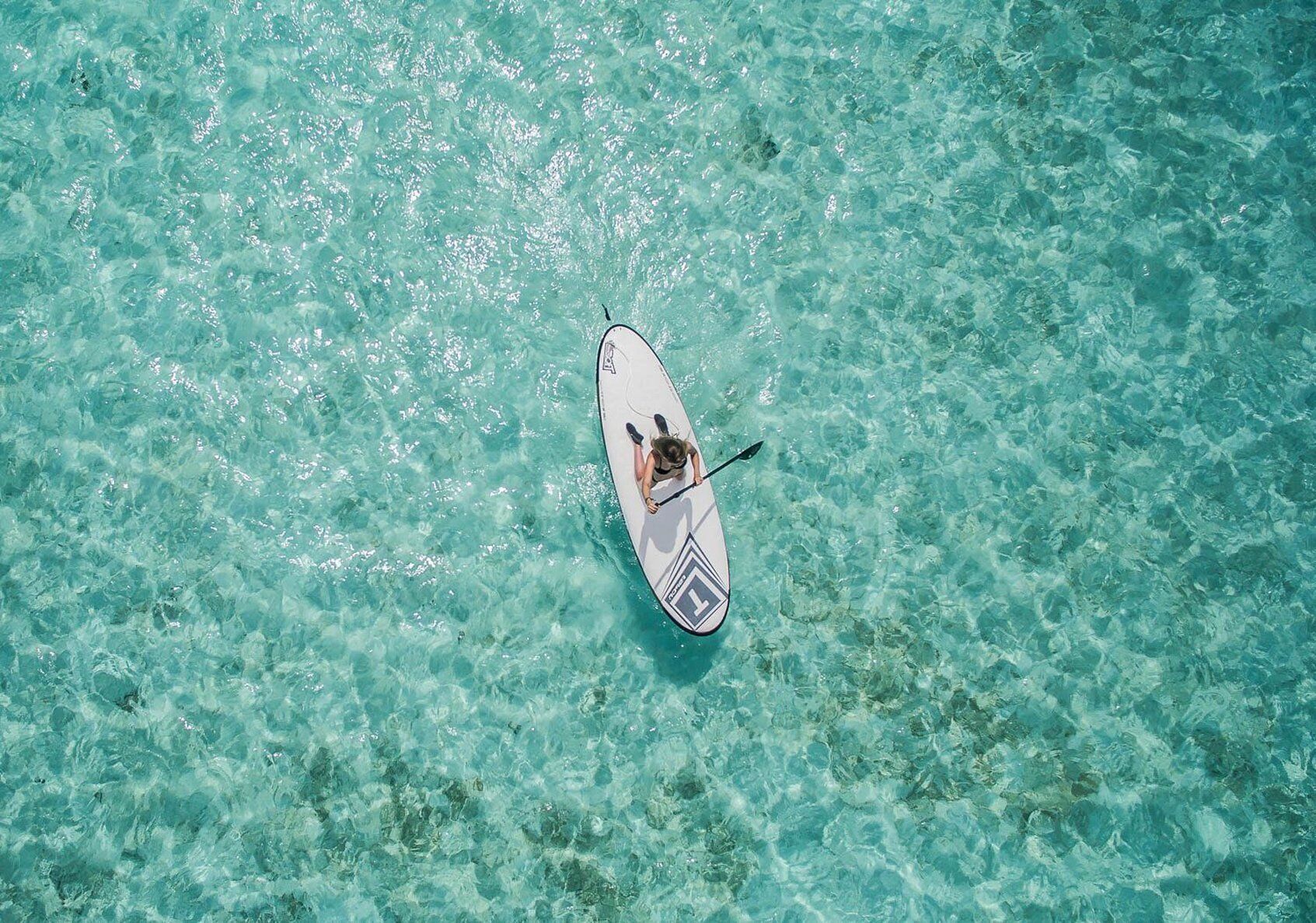 An aerial view of a clear turquoise sea with a single paddleboard and a person lying on it.