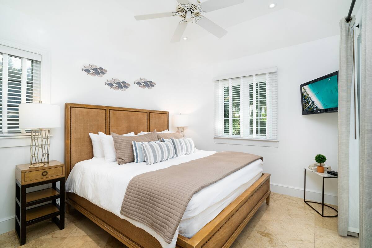 A bedroom with a wooden bed, white bedding, a nightstand, a TV on the wall, and a window with blinds.