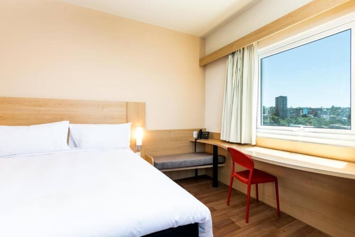 A hotel room with a bed, a desk with a chair, a window with curtains, and a view of a cityscape.