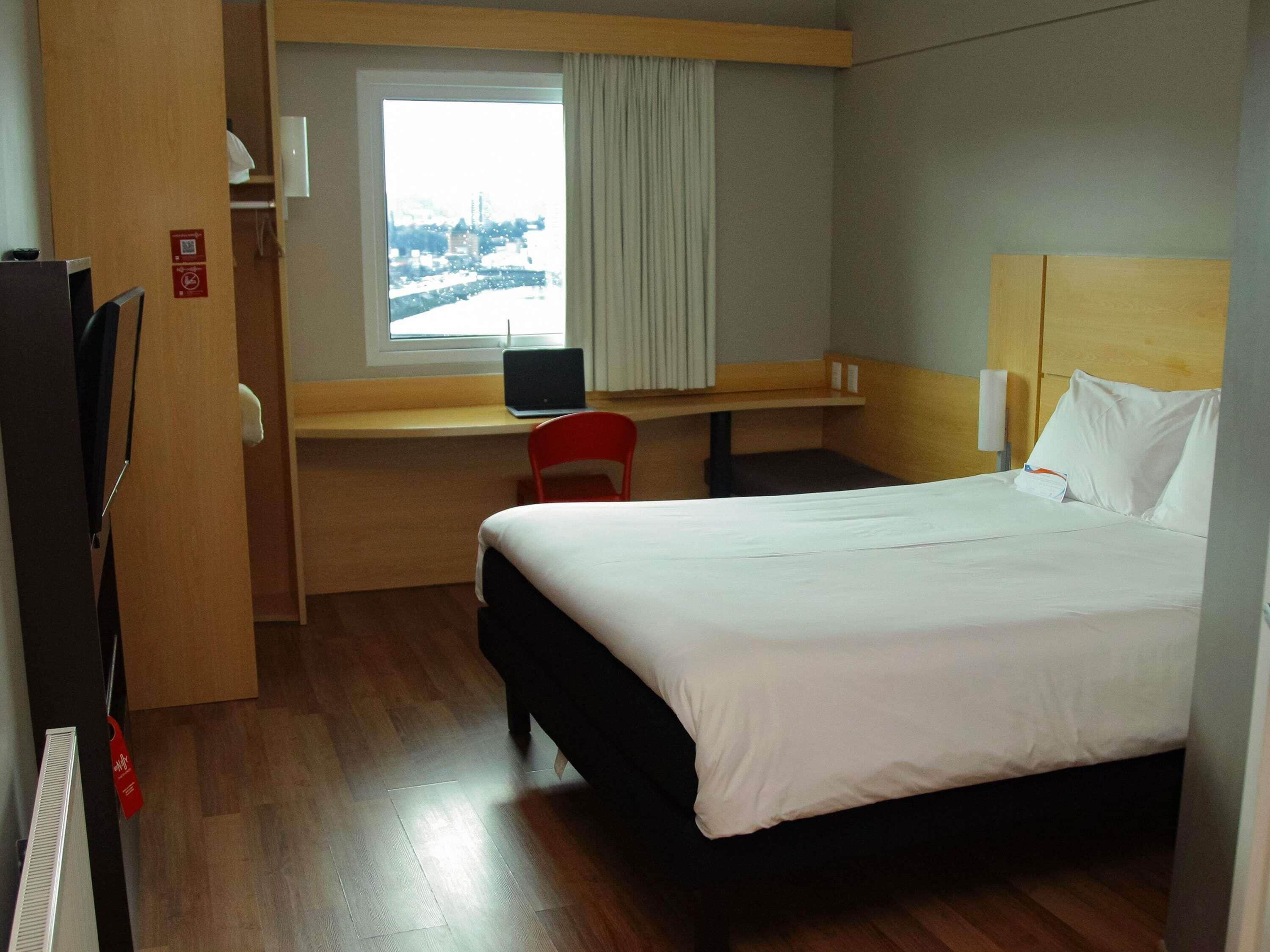 A hotel room with a bed, a desk with a laptop, a chair, and a window with a view.