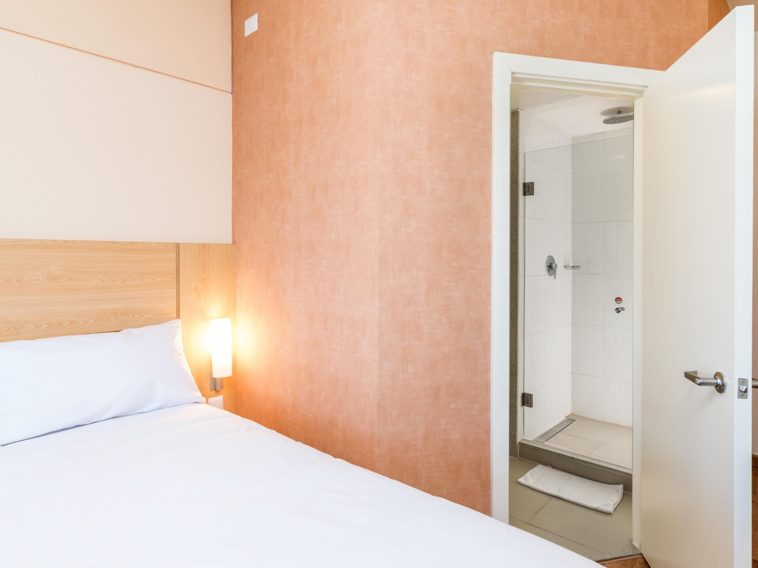 Hotelkamer met een bed, nachtkastje met lamp, en een open deur naar een badkamer met douche en toilet.