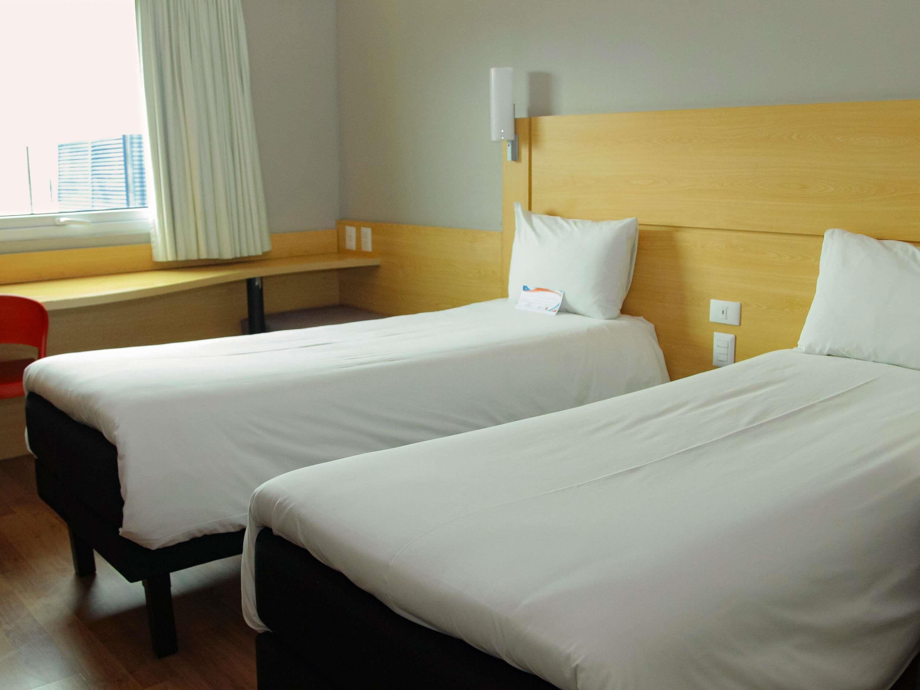 Een hotelkamer met twee enkele bedden, een houten hoofdbord, een bureau en een raam met gordijnen.
