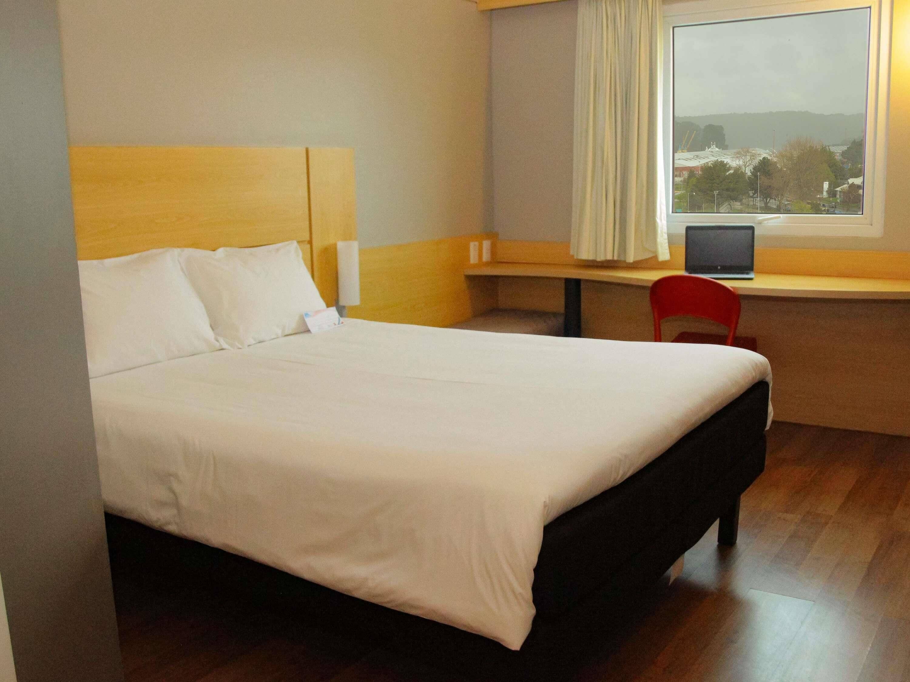 A hotel room with a large bed, a desk with a chair, a laptop, and a window with a view.