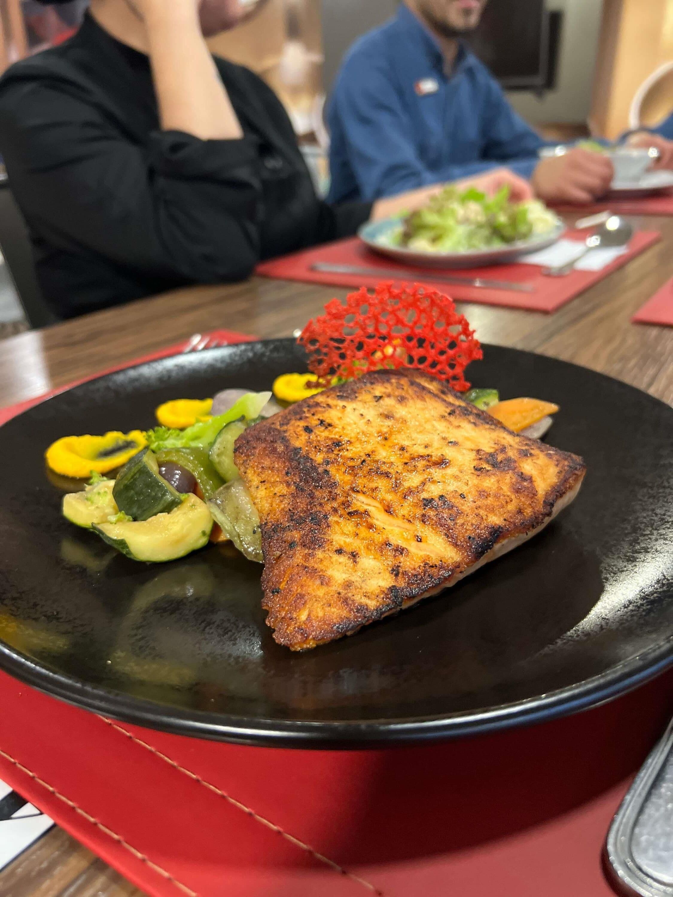 Un plato con pescado a la parrilla, verduras y un detalle decorativo rojo.