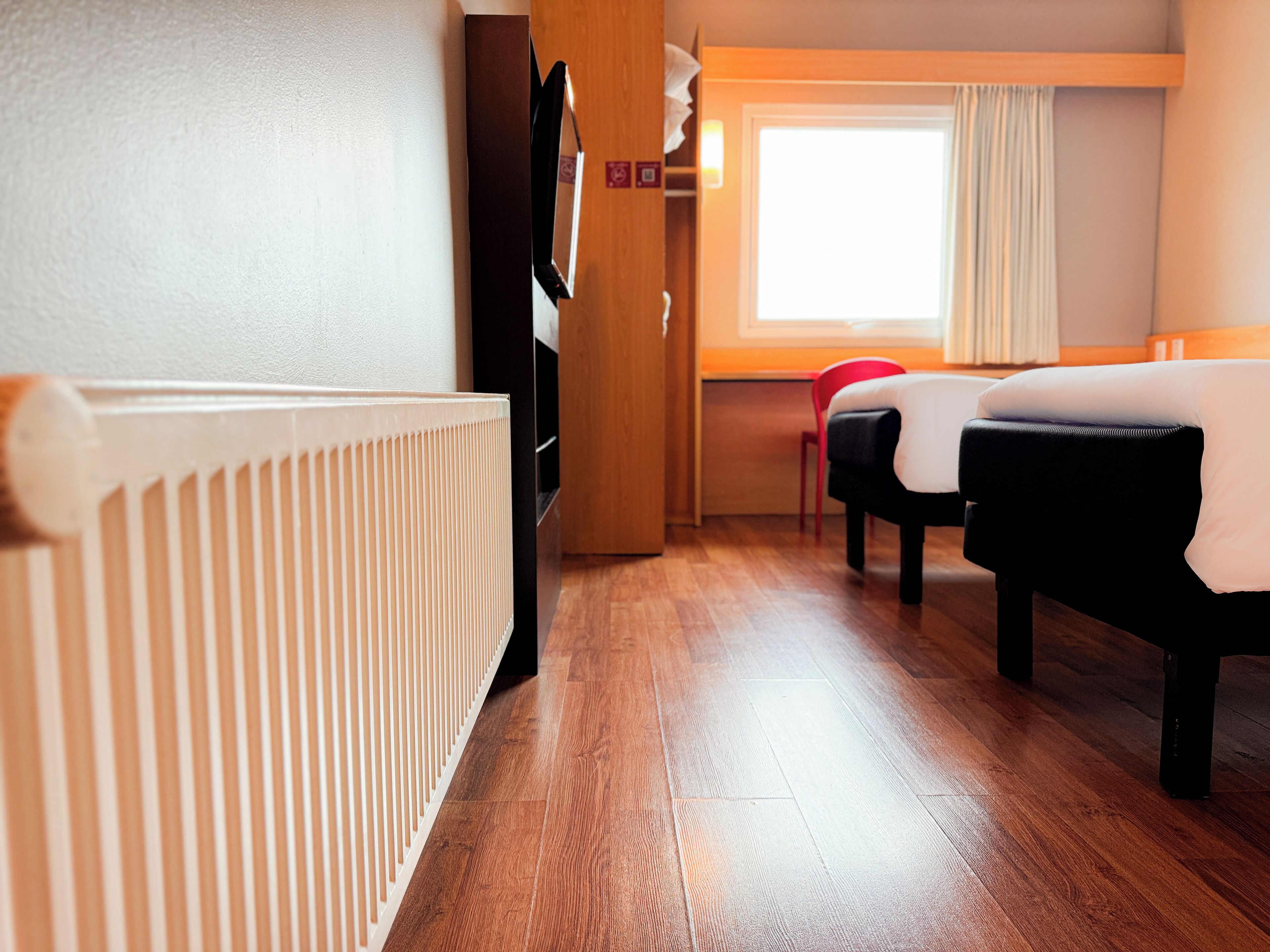 A hotel room with a wooden floor, a radiator, a bed with a white duvet, a red chair, and a window with curtains.