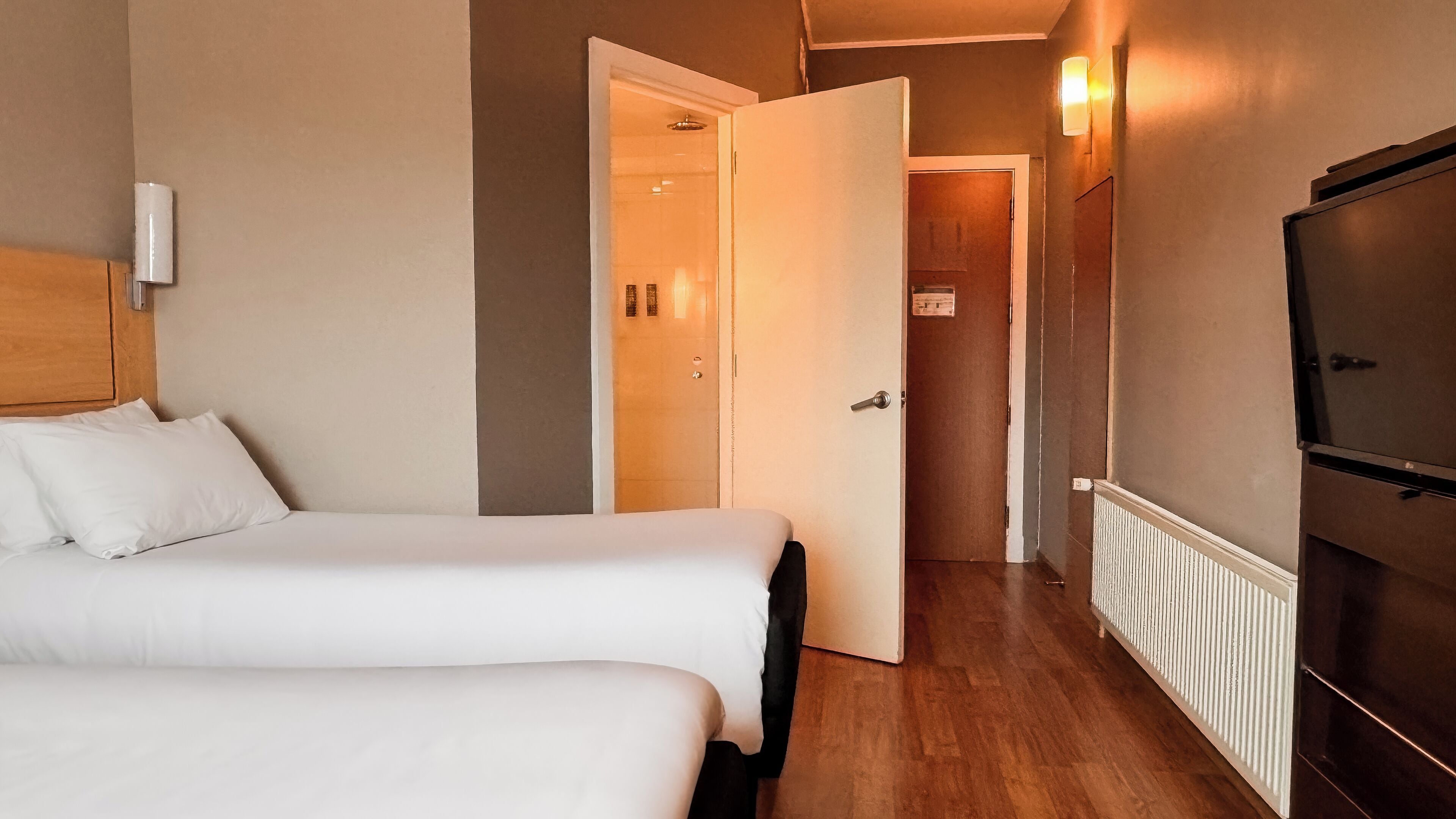 Une chambre d’hôtel avec deux lits, une armoire en bois, un téléviseur et un couloir menant à une salle de bain.