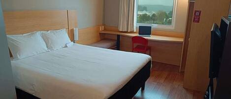 A hotel room with a bed, a desk with a laptop, a chair, and a window with a view.