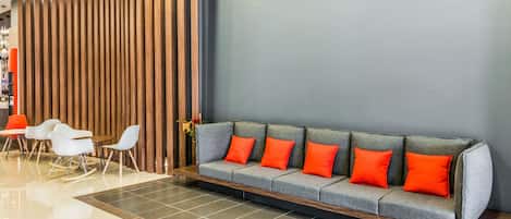 Un área de recepción moderna con un sofá gris, cojines naranjas y sillas blancas.