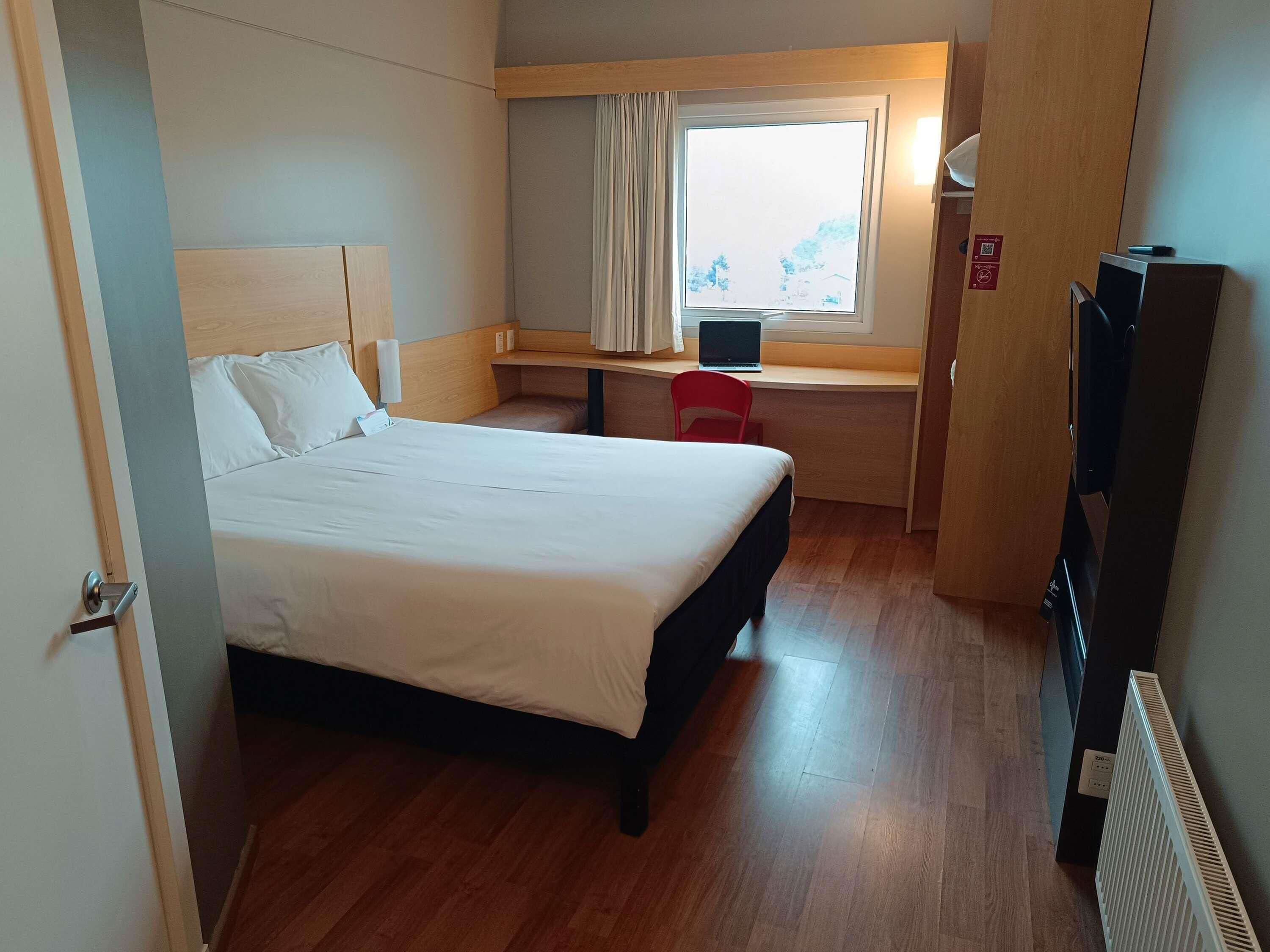 Une chambre d’hôtel avec un lit, un bureau avec un ordinateur portable, une chaise rouge, une télévision et une fenêtre avec des rideaux.