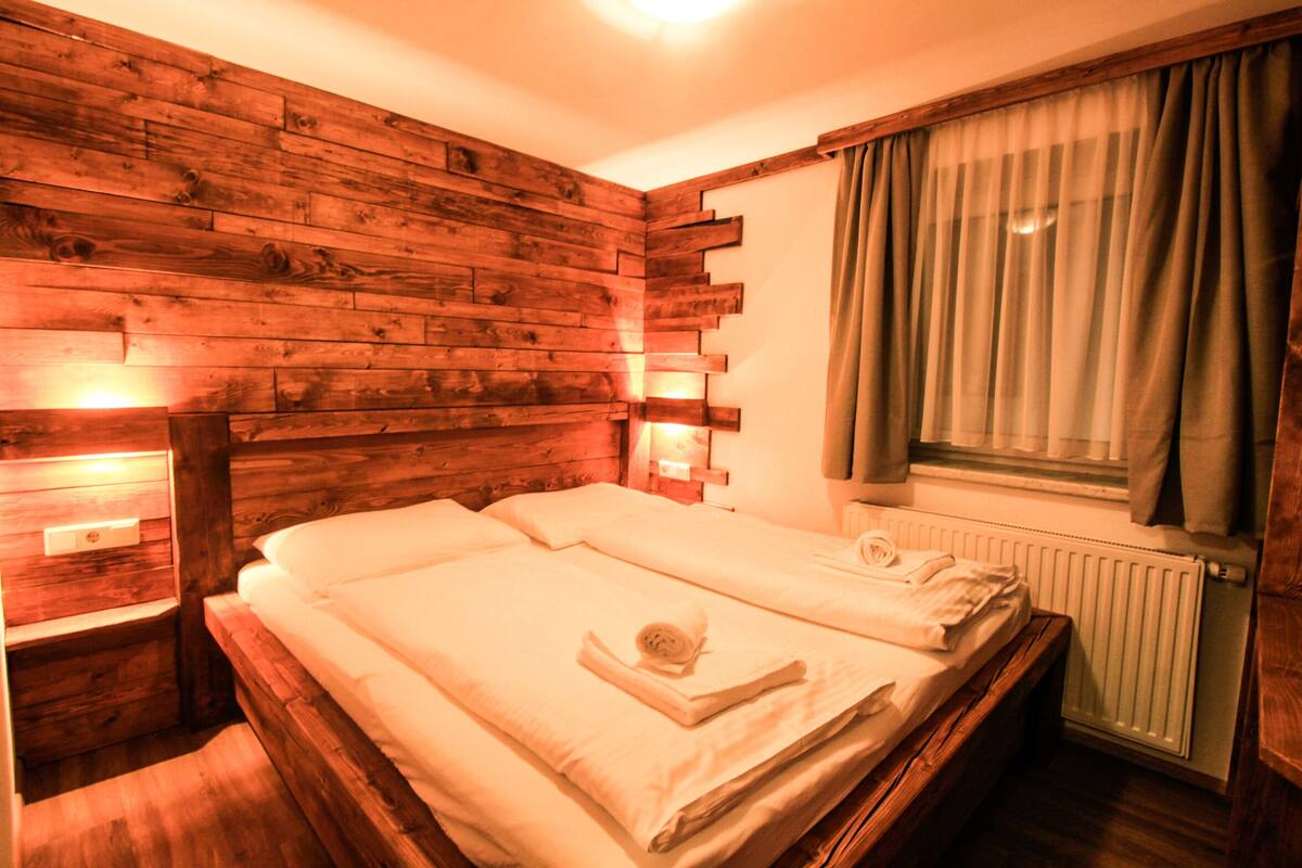 A wooden-paneled room with a bed, a nightstand, and a window with curtains.