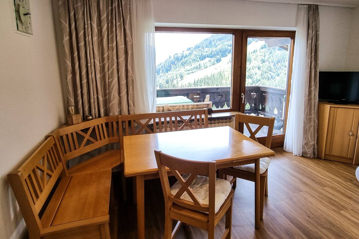 A wooden dining table with chairs, a television on a cabinet, and a view of a mountainous landscape through a large window.
