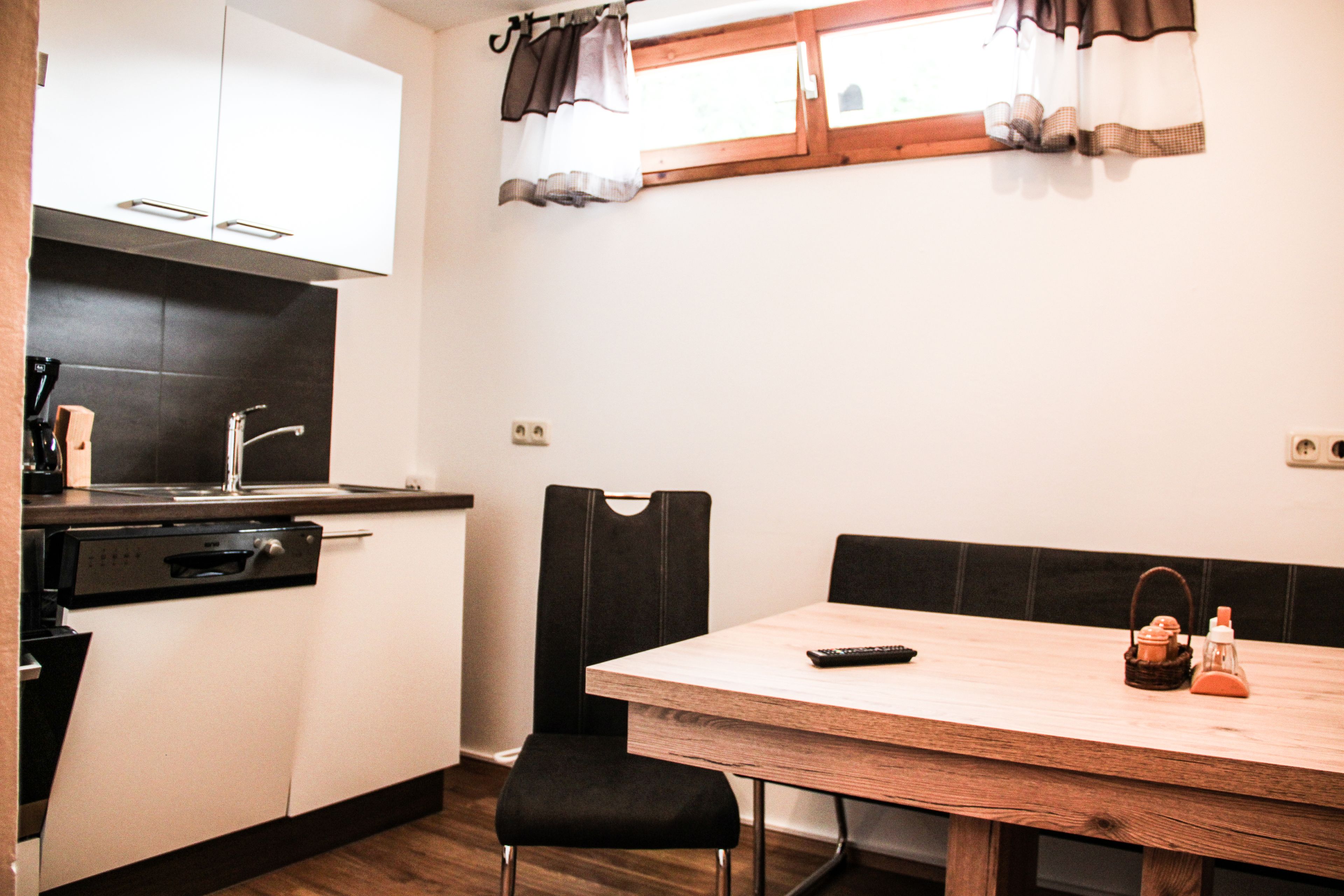 Een compacte keuken met een vaatwasser, een spoelbak en een raam met gordijnen. Er staat een houten tafel met een stoel en een afstandsbediening.