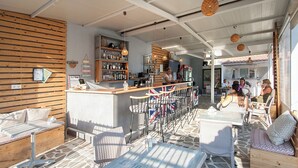 A bar area with wooden slats, a counter, and bar stools. There are tables and chairs for seating. - Dream House (Skiathos)