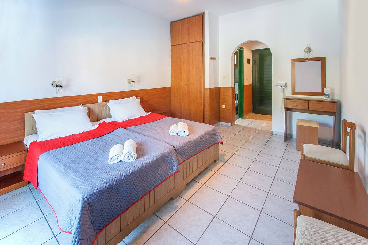 A hotel room with two beds, a wooden wardrobe, a small desk with a mirror, and a bathroom visible through an archway.
