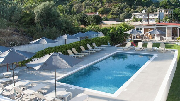 A rectangular swimming pool surrounded by white lounge chairs and umbrellas, with a view of a hillside and buildings in the background. - Dream House (Skiathos)