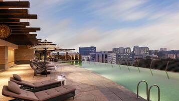 An outdoor pool area with lounge chairs and umbrellas, a city skyline in the background, and a clear sky.