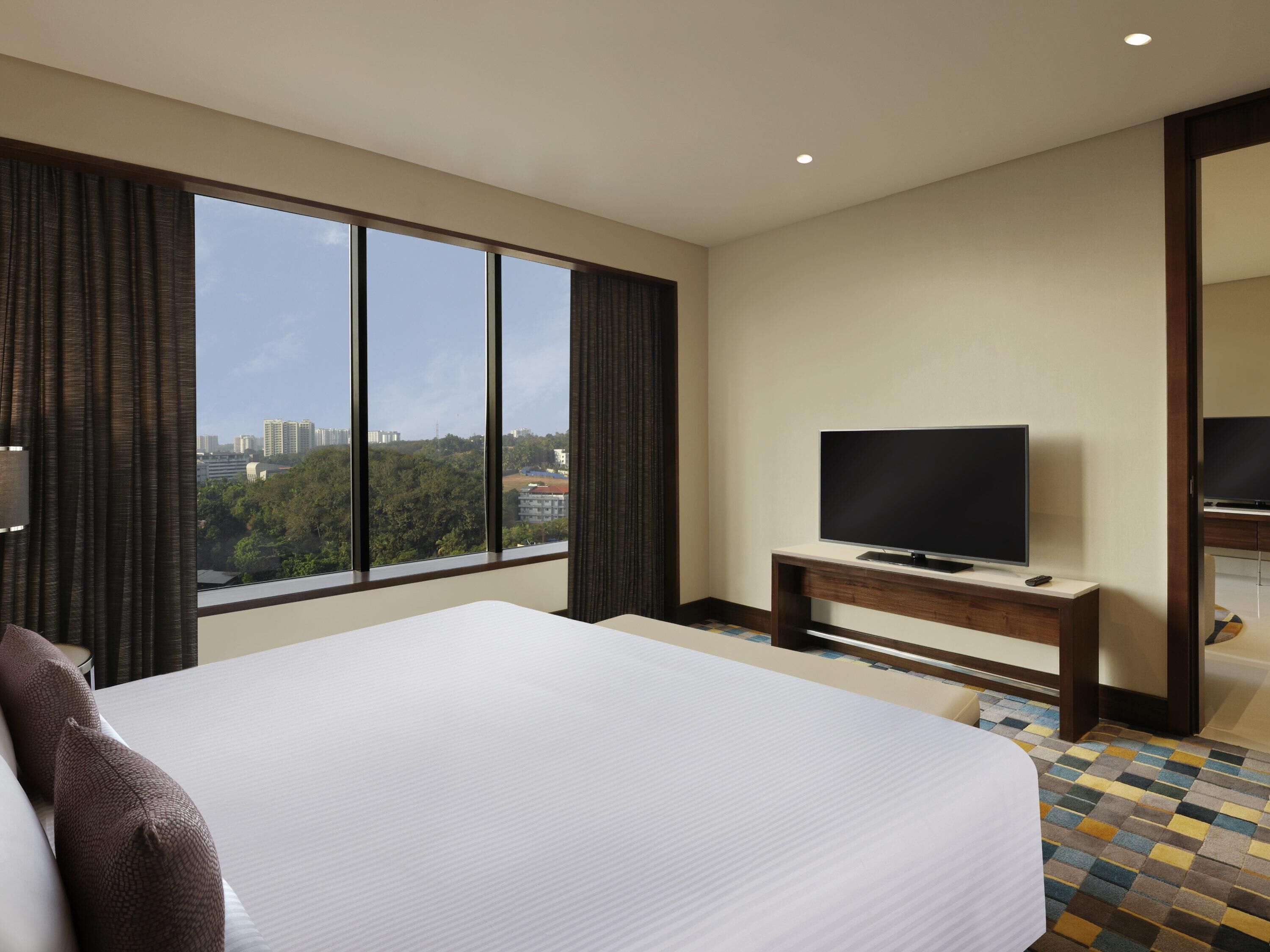 A hotel room with a large bed, a television on a wooden stand, and a view of the city through large windows.