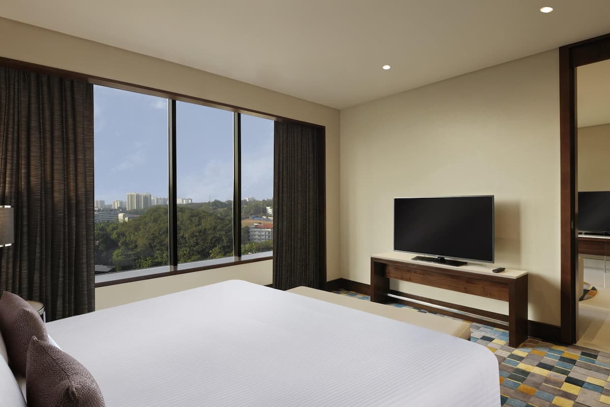 A hotel room with a large bed, a television on a wooden stand, and a view of the city through large windows.