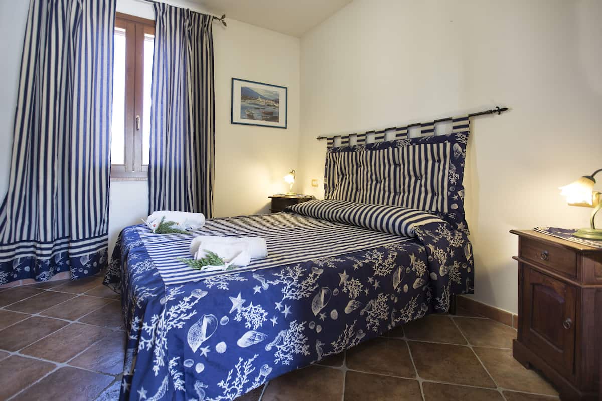 Un dormitorio con cama, cortinas, mesita de noche, lámpara, un cuadro enmarco y un armario de madera.