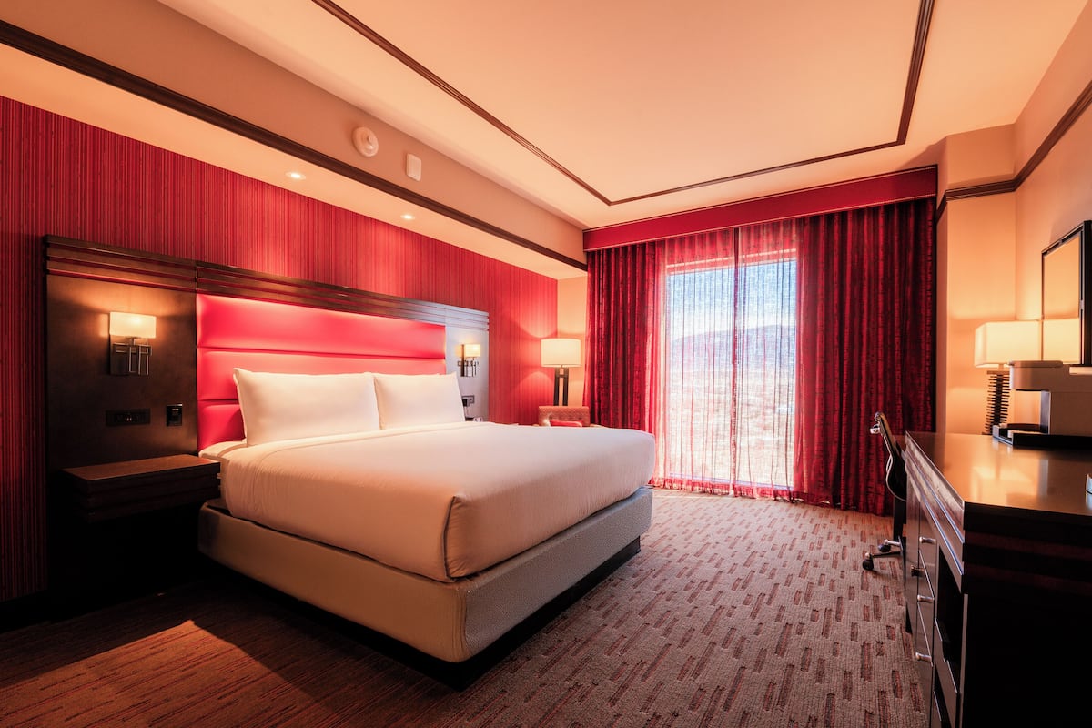 A hotel room with a large bed, a nightstand, a desk, and a window with red curtains.