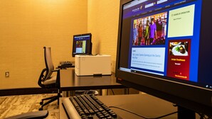 Una habitación de hotel con un escritorio, un monitor de computadora mostrando una página web, una impresora y una silla.