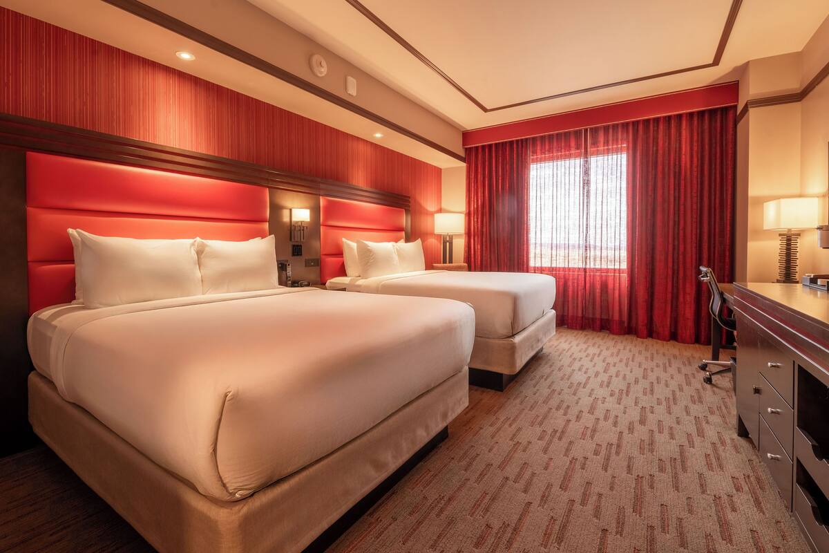 A hotel room with two beds, a desk, and a large window with red curtains.