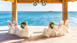 A wooden pergola with a view of the sea, four chairs with white covers and pink ribbons, and two green wreaths. - Serene by the Sea (Savaneta)