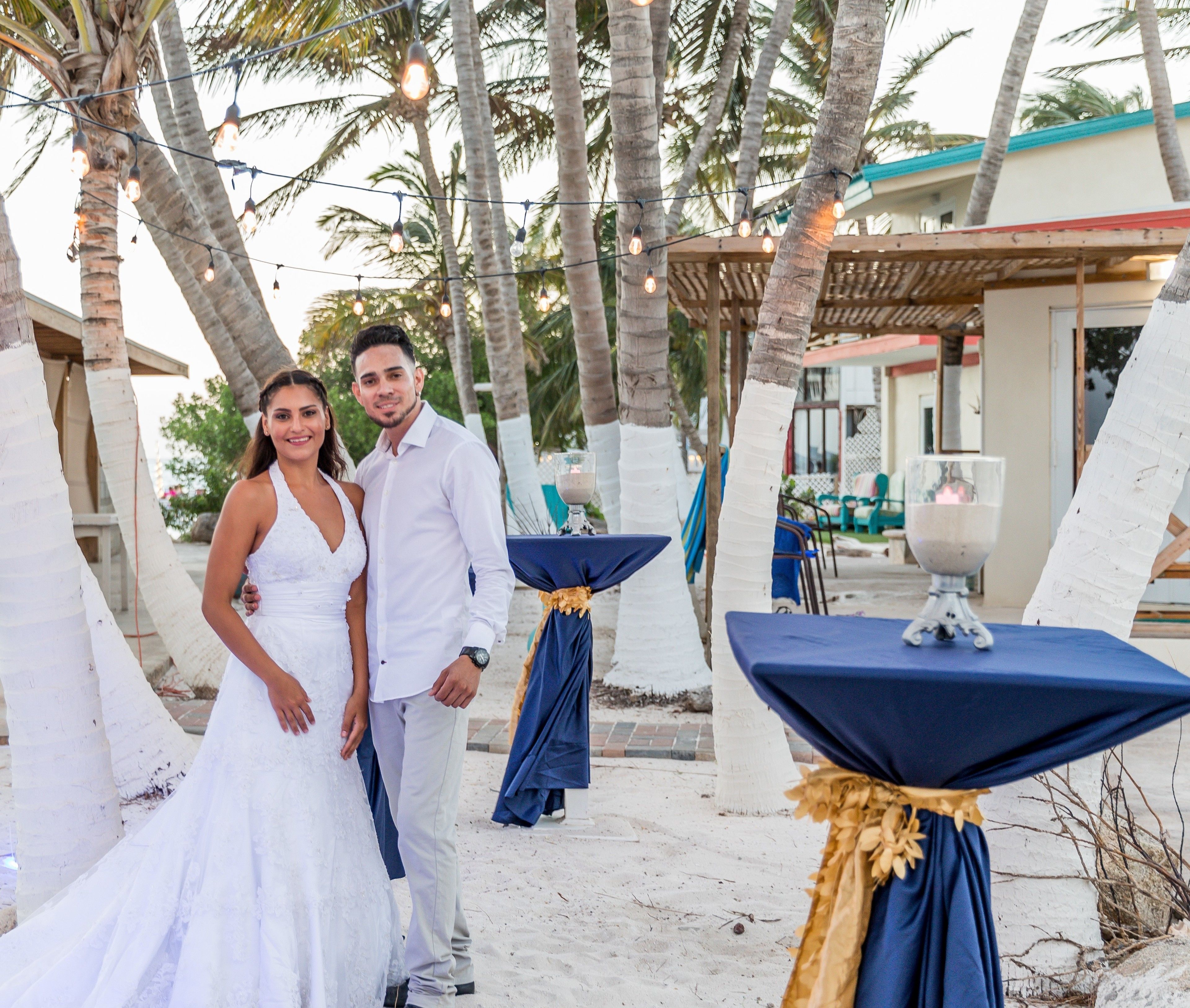 A wedding ceremony outdoors with a sandy area, palm trees, and a building in the background.